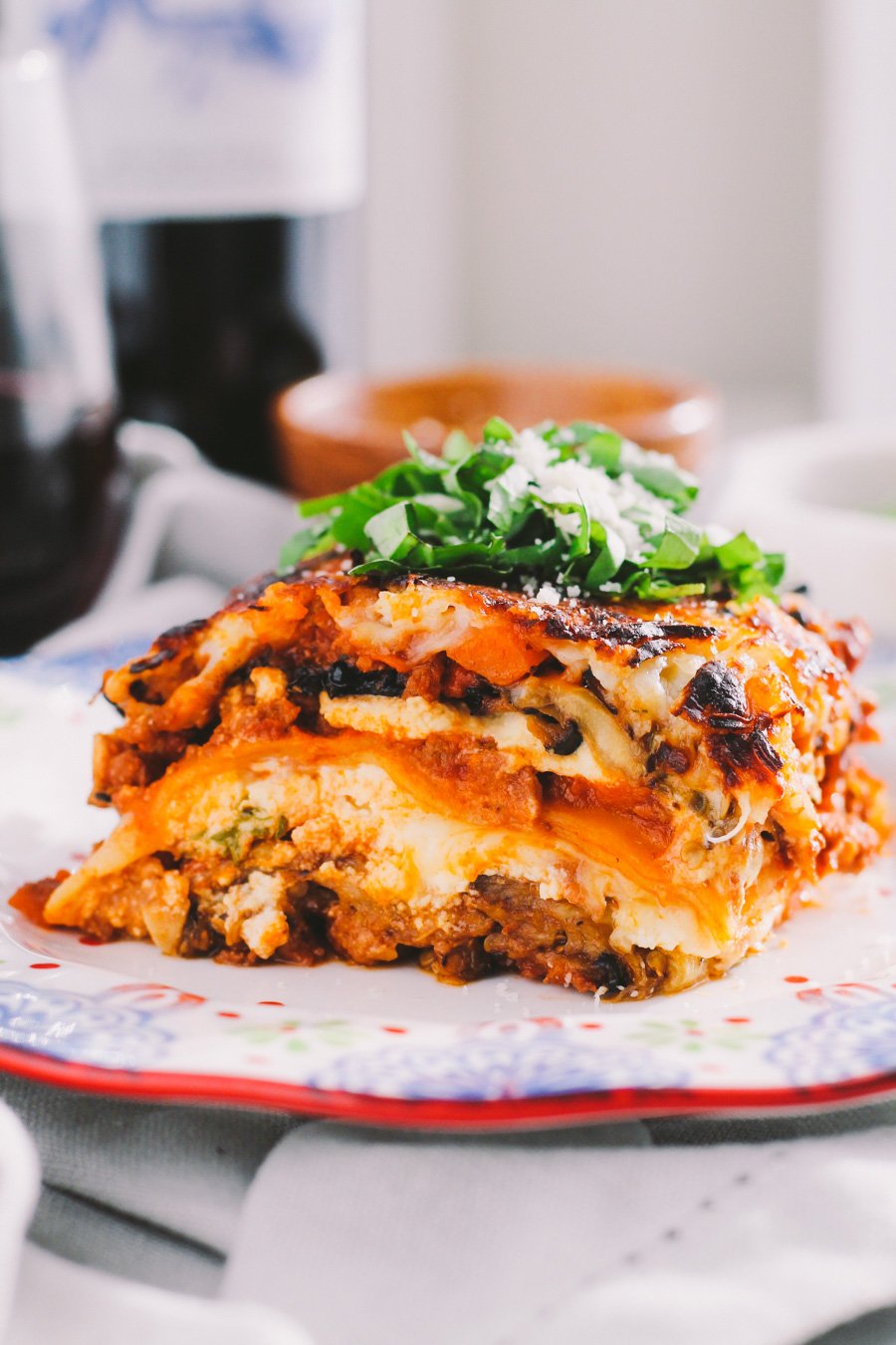 A side angle shot of eggplant lasagna served on a colorful patterned plate. The plate sits atop a white and gray marble surface with a glass of red wine and a small wooden pinch bowl filled with grated parmesan cheese.