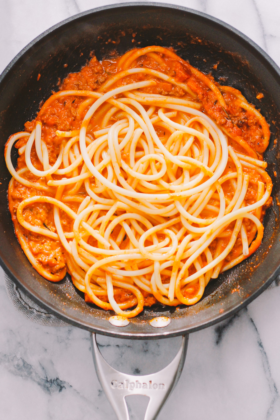 A macro close up shot of bucatini pasta tossed with ground turkey bolognese sauce inside of a skillet. A pair of metal tongs are used to lift & toss the pasta in the skillet.