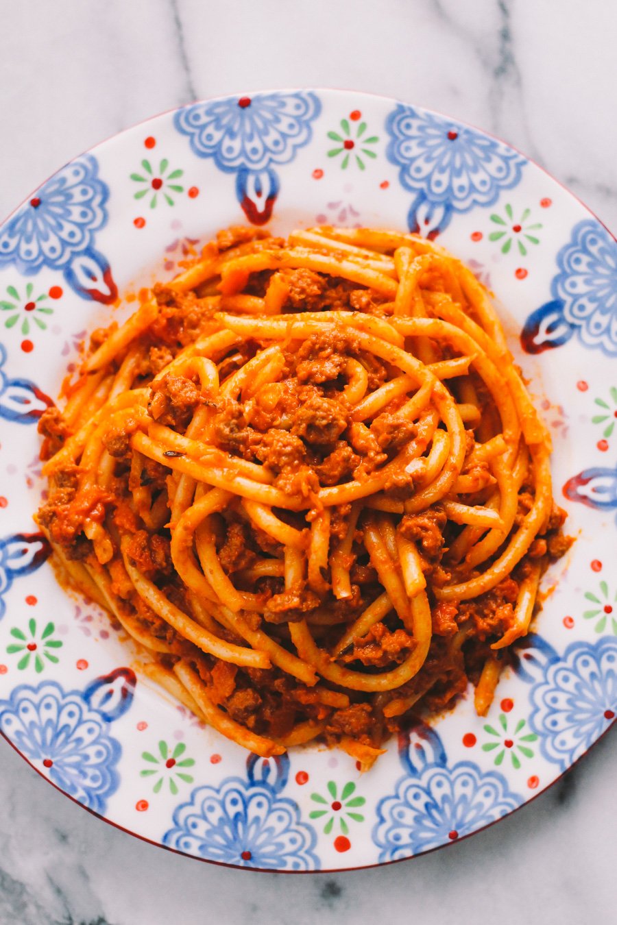 An overhead shot of ground turkey bolognese sauce served with bucatini atop a colorful patterned plate. The plate sits atop a white and gray marble surface.
