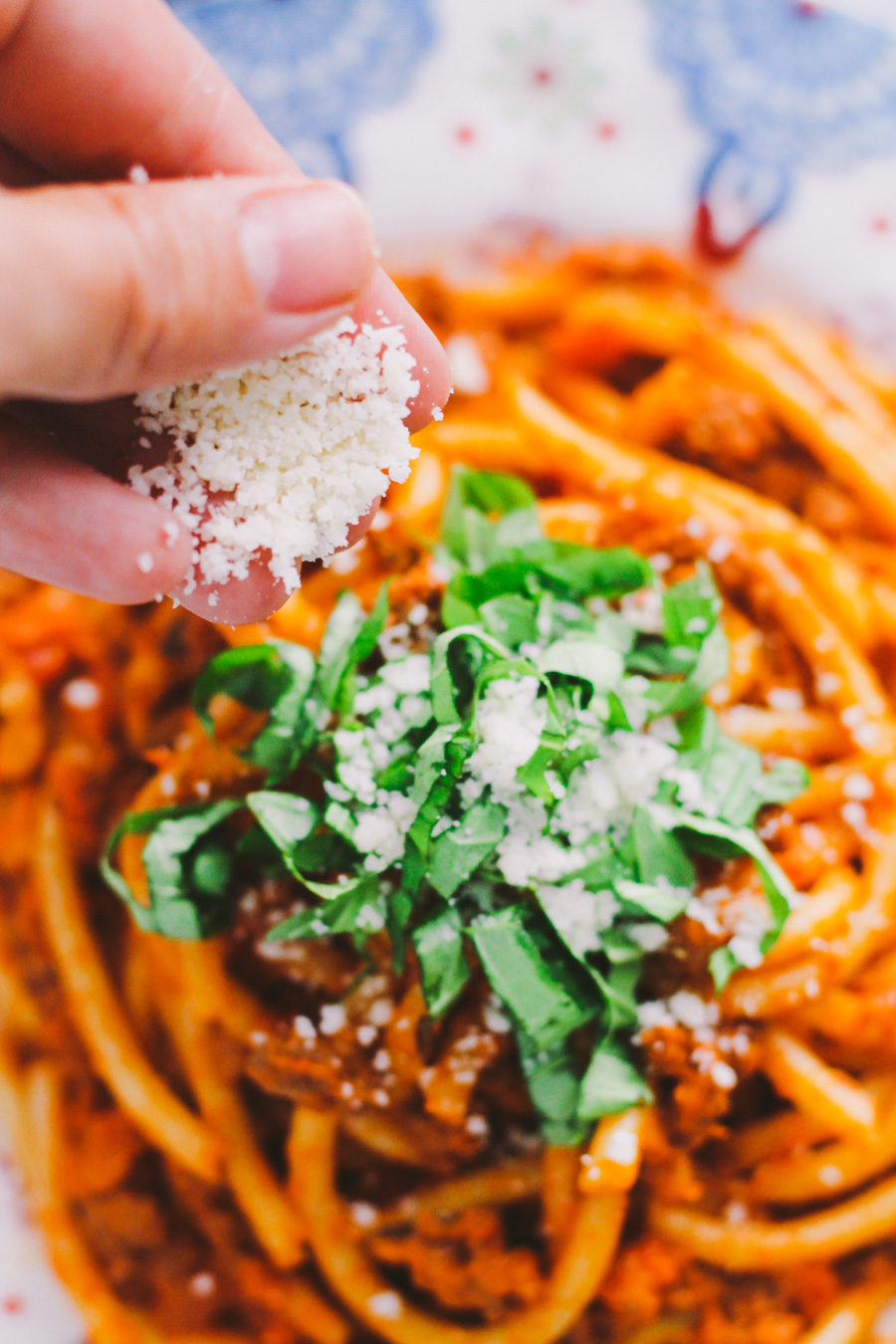 An overhead shot of ground turkey bolognese sauce served with bucatini atop a colorful patterned plate. The pasta has been garnished with fresh chopped basil. A woman's hand works to sprinkle fresh grated parmesan cheese overtop of the pasta.