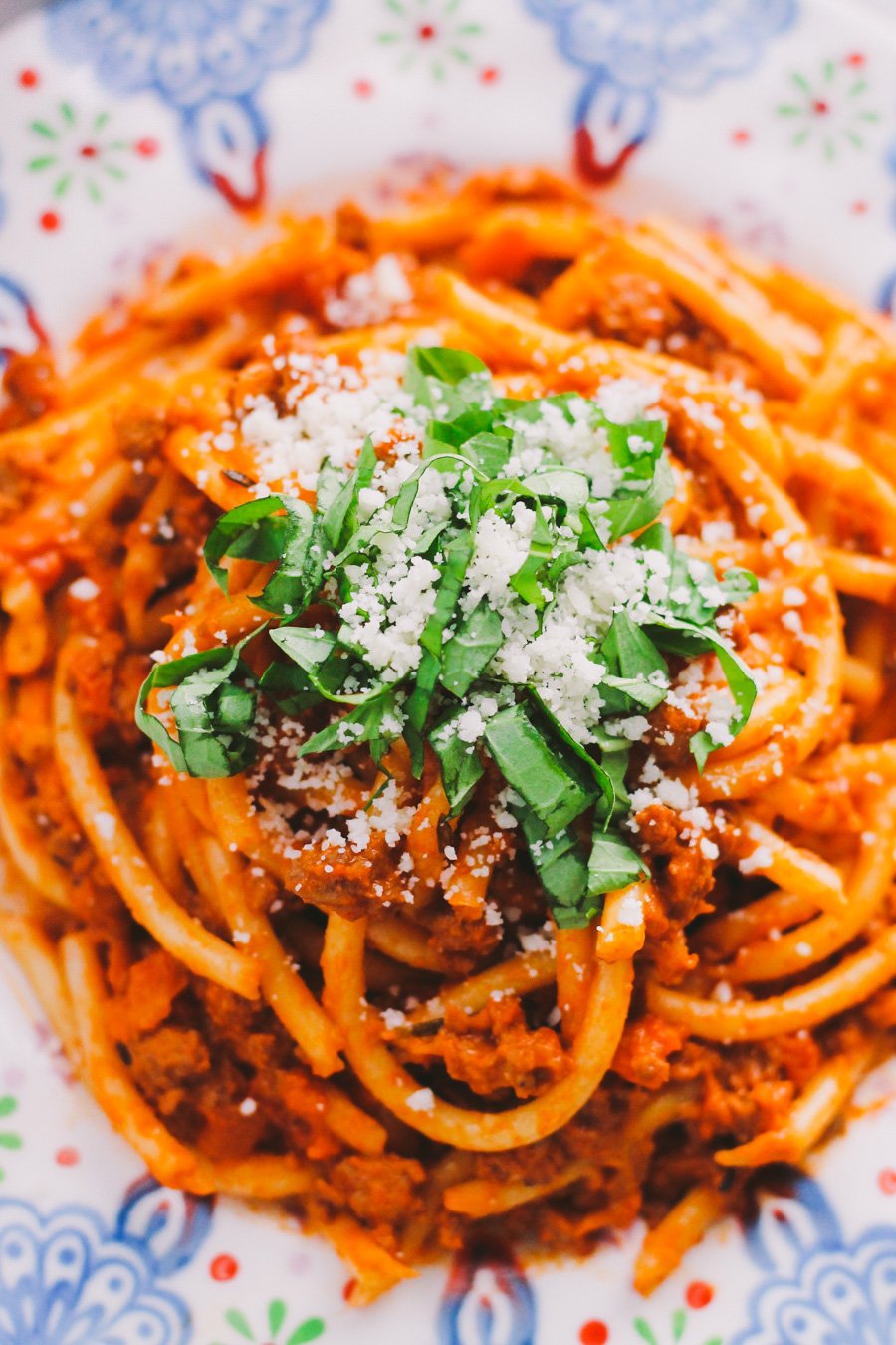 An overhead and close up shot of ground turkey bolognese sauce served with bucatini atop a colorful patterned plate. The plate sits atop a white and gray marble surface. The pasta has been garnished with fresh chopped basil and fresh grated parmesan cheese.