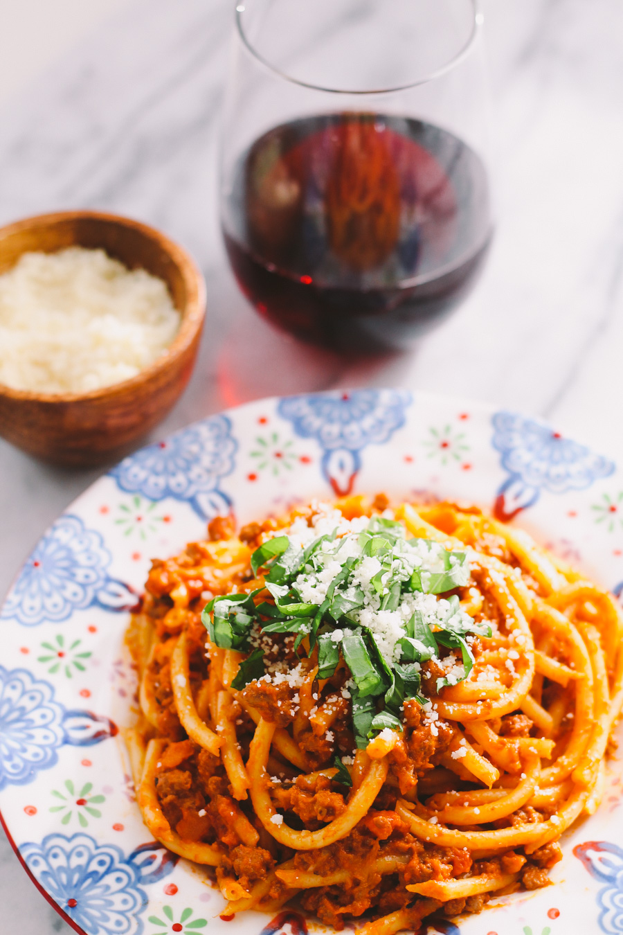 An overhead and close up shot of ground turkey bolognese sauce served with bucatini atop a colorful patterned plate. The plate sits atop a white and gray marble surface. The pasta has been garnished with fresh chopped basil and fresh grated parmesan cheese. A small wooden pinch bowl filled with grated parmesan and a glass of red wine rest alongside the plate.