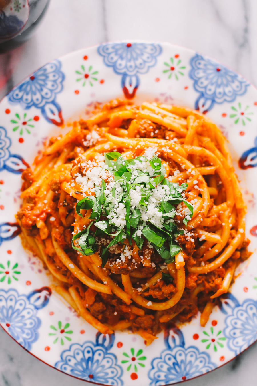 Turkey bolognese tossed with bucatini is served atop a patterned plate. The pasta is garnished with grated parmesan cheese and fresh sliced basil.