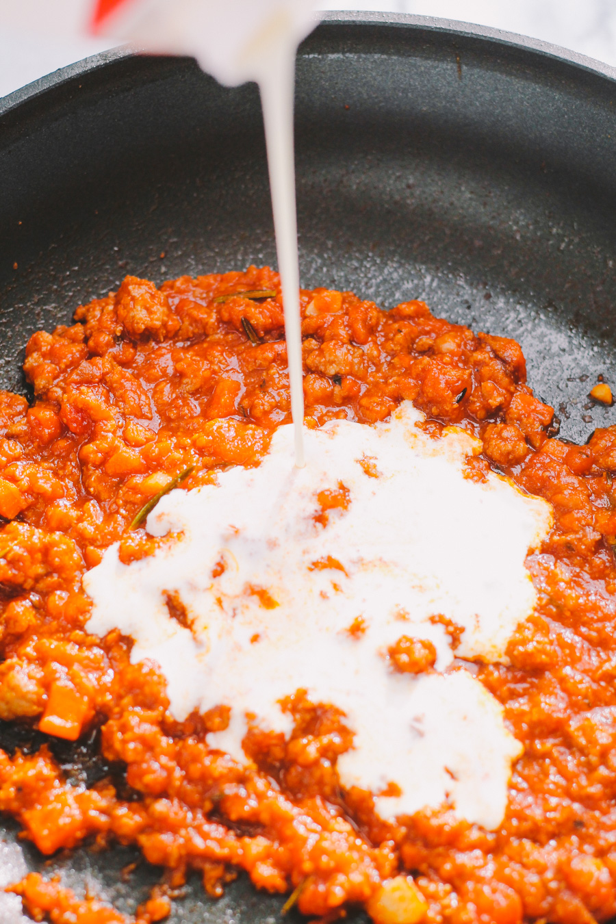 An overhead shot of heavy cream being added to a ground turkey pasta fauxlognese sauce inside of a skillet.