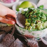 A woman's hand holds a blue corn tortilla chip as she scoops up a chipful of guacamole inside of a colorful ceramic bowl. The bowl is surrounded by guacamole ingredients and more blue corn tortilla chips for dipping.