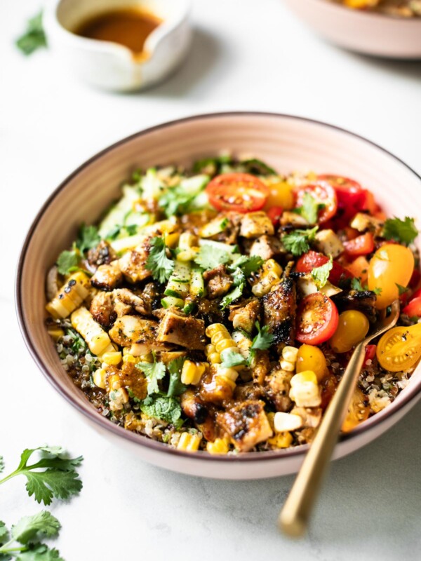 Side angle of a summer glow bowl in a shallow pink bowl: a bed of quinoa topped with sliced tomatoes, diced red bell pepper, grilled corn kernels & shredded cucumber. The bowl has a small gold fork in it. It sits atop a white marble surface, next to a small dish of curry vinaigrette & a couple of summer tomatoes.