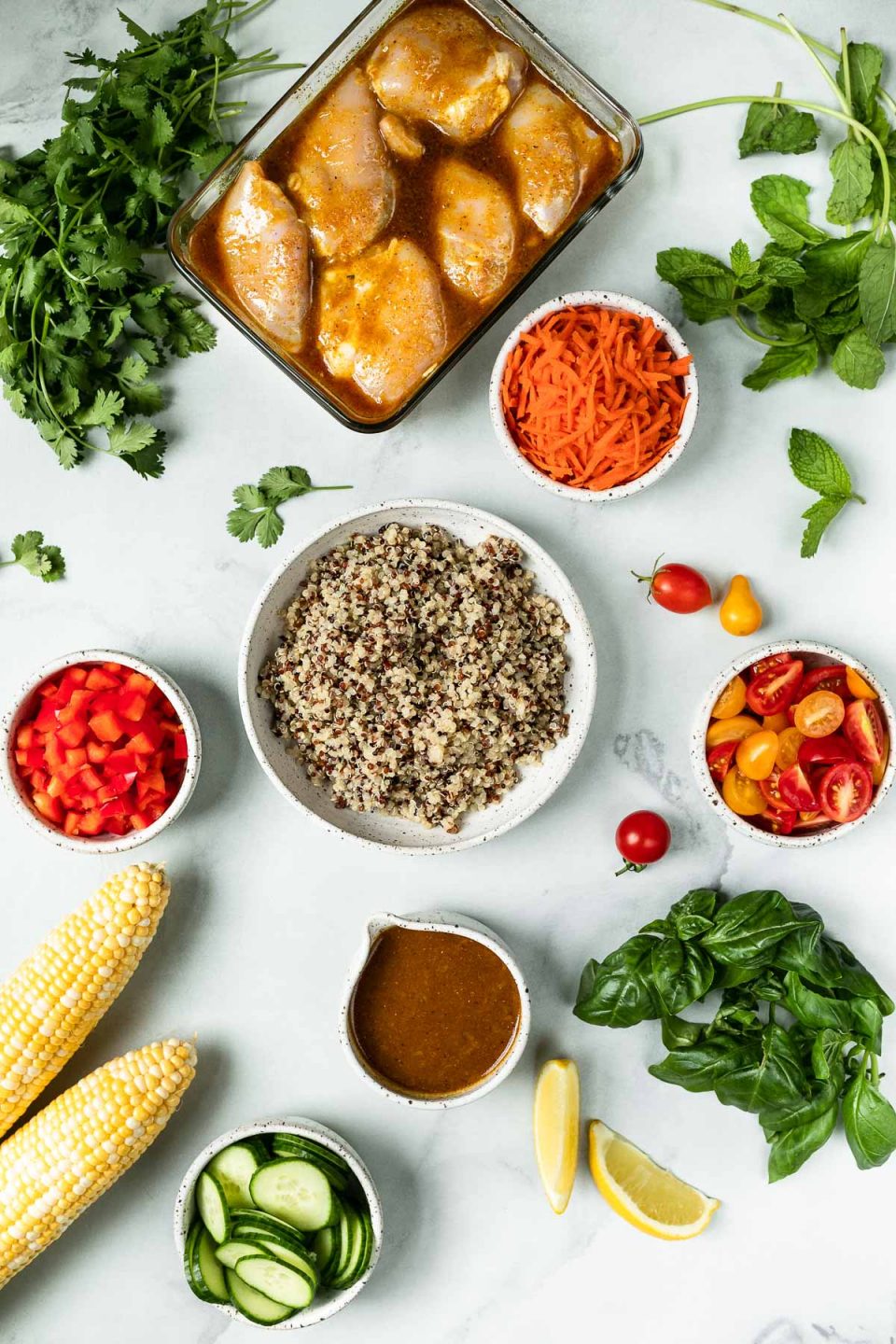 Glow bowls components arranged on a white marble surface: marinated chicken thighs, grated carrot, quinoa, diced bell pepper, cucumber, corn, & fresh herbs (cilantro, mint & basil).