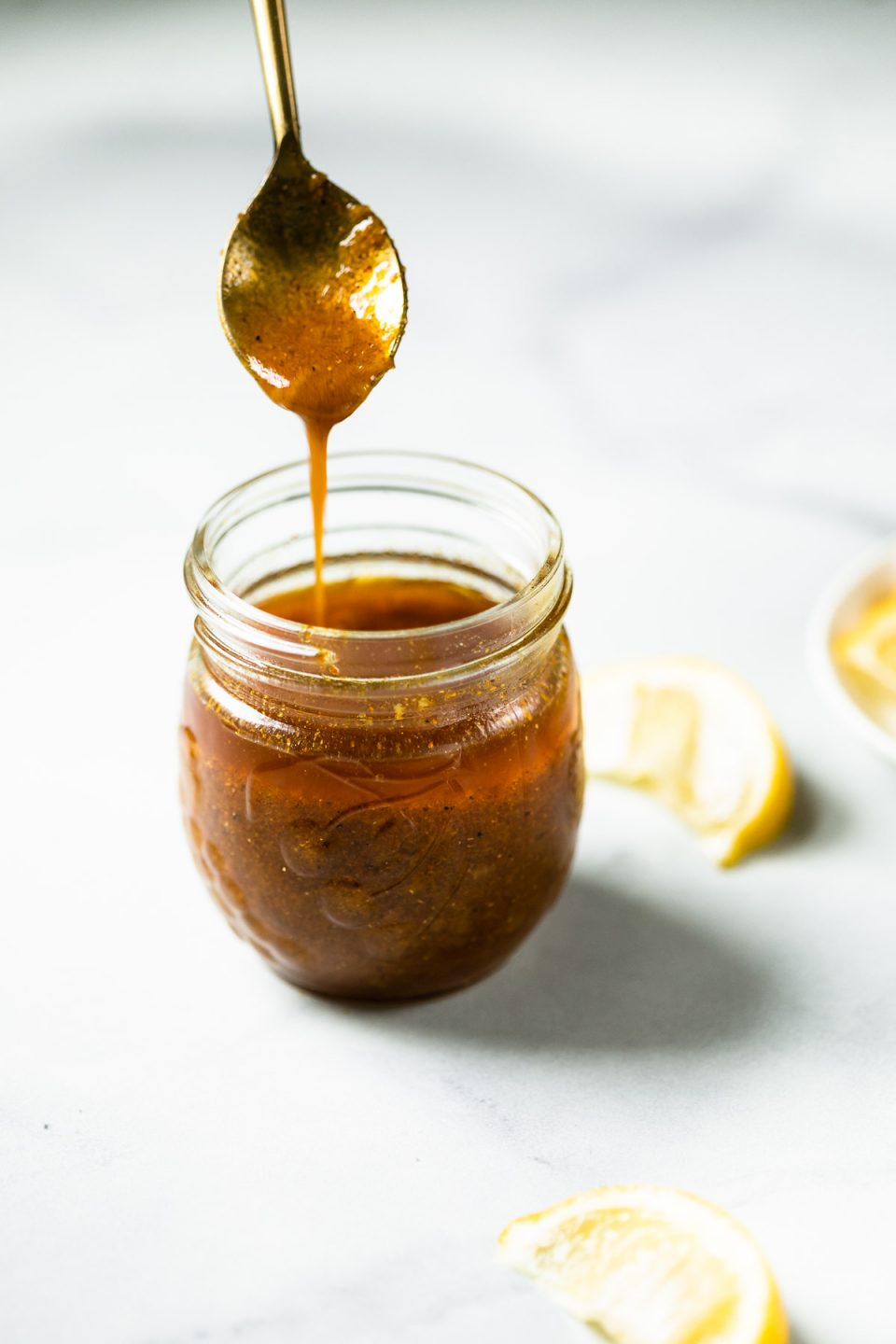 Curry vinaigrette in a small glass jar, with a golden spoon drizzling some of the vinaigrette into the jar. The jar is surrounded by sliced lemon wedges.