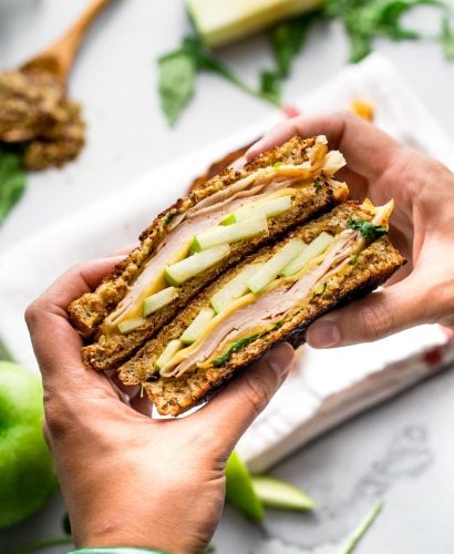 A woman's hand holds two halves stacked of an Autumn Grilled Cheese Sandwich. This apple grilled cheese contains gouda cheese, granny smith apples, whole grain mustard, smoked turkey, & arugula. Out of focus & in the background there is a white linen napkin, pieces of granny smith apple, a wooden spoon filled with mustard, & pieces of arugula resting on a grey plaster surface.