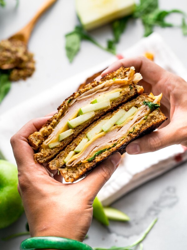 A woman's hand holds two halves stacked of an Autumn Grilled Cheese Sandwich. This apple grilled cheese contains gouda cheese, granny smith apples, whole grain mustard, smoked turkey, & arugula. Out of focus & in the background there is a white linen napkin, pieces of granny smith apple, a wooden spoon filled with mustard, & pieces of arugula resting on a grey plaster surface.