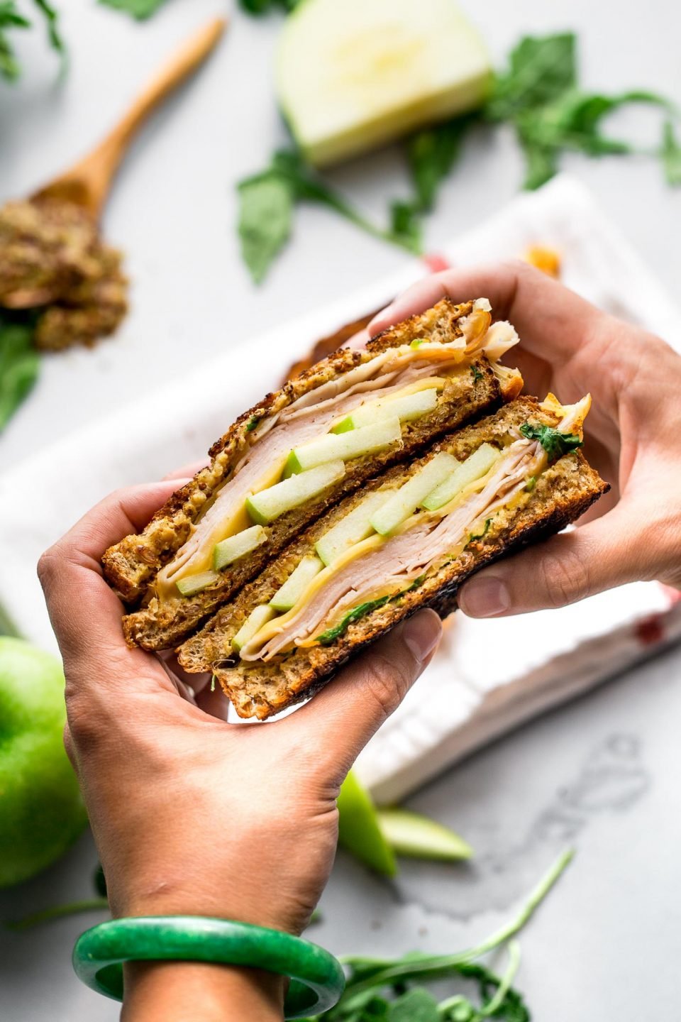 A woman's hand holds two halves stacked of an Autumn Grilled Cheese Sandwich. This apple grilled cheese contains gouda cheese, granny smith apples, whole grain mustard, smoked turkey, & arugula. Out of focus & in the background there is a white linen napkin, pieces of granny smith apple, a wooden spoon filled with mustard, & pieces of arugula resting on a grey plaster surface.