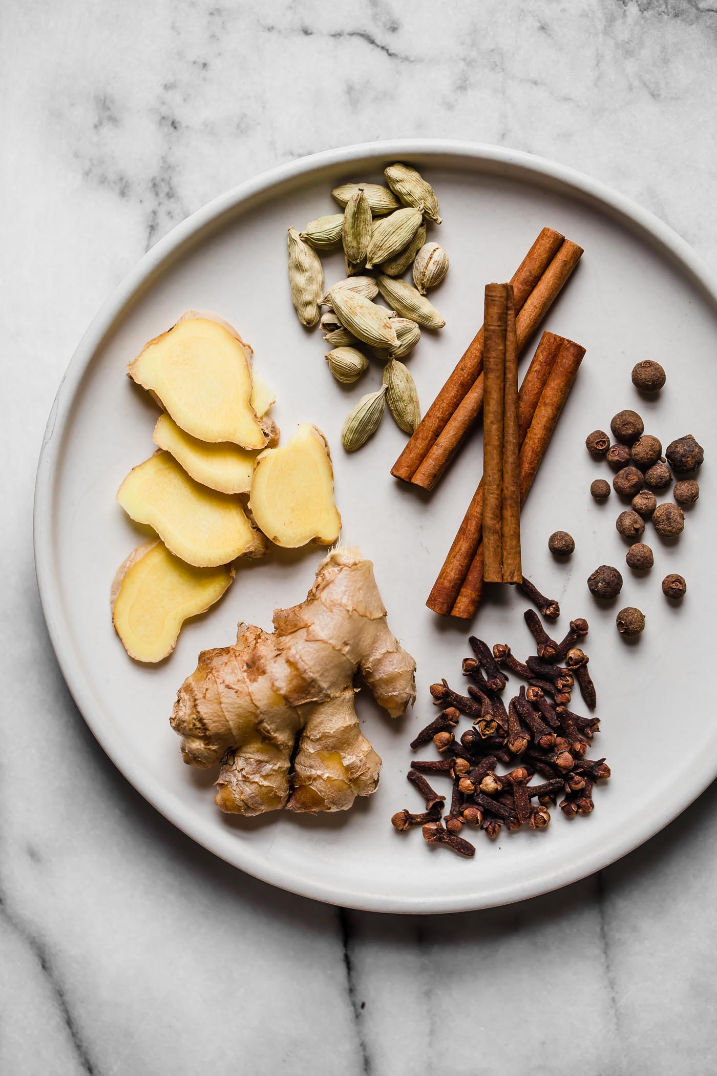 Homemade chai spice ingredients arranged on a white ceramic plate: cinnamon, cardamom, ginger, allspice, cloves, & nutmeg. The plate sits atop a white & gray marble surface.