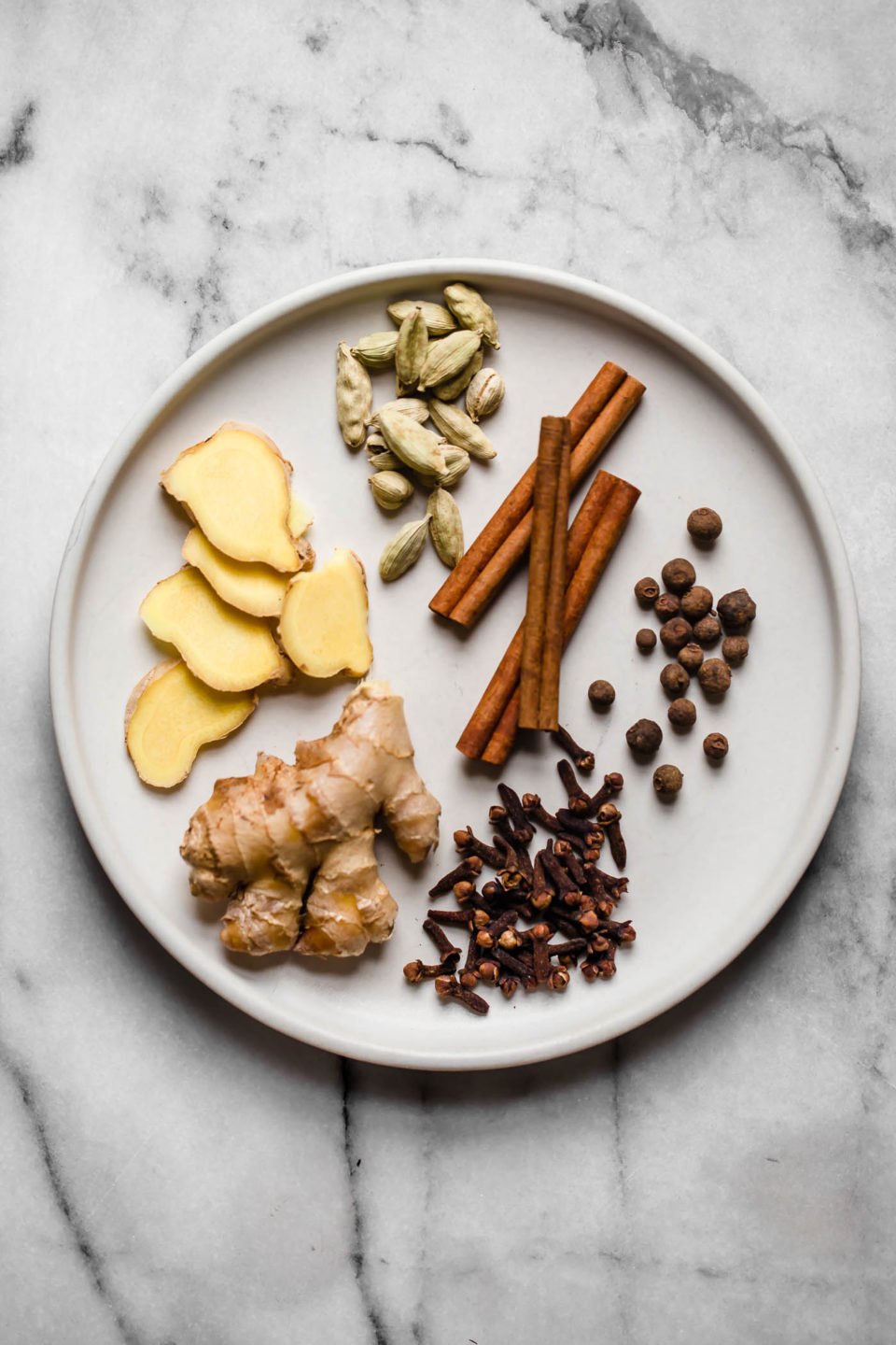Homemade chai spice ingredients arranged on a white ceramic plate: cinnamon, cardamom, ginger, allspice, cloves, & nutmeg. The plate sits atop a white & gray marble surface.