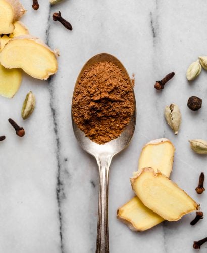 A silver spoon rests on a white & gray marble surface filled with a spoonful of ground chai spice mix. Surrounding the spoon are several raw forms of the spices that go into this homemade chai spice mix.
