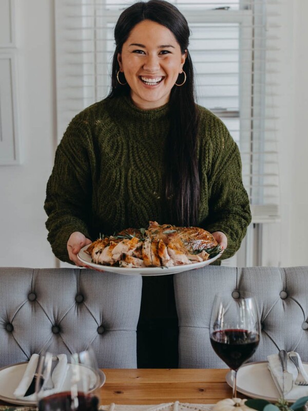 Dark-haired woman in a green sweater holds a plate of carved turkey, in front of a festive tablescape & a full Thanksgiving dinner spread.