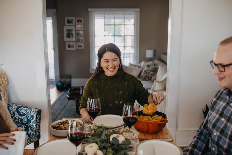 A dark-haired woman in a green sweater seated at the head of a Thanksgiving table. She is serving herself no-stir pumpkin risotto from a pumpkin-shaped Dutch oven.