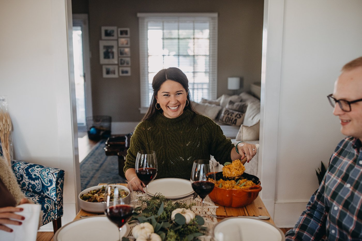 A dark-haired woman in a green sweater seated at the head of a Thanksgiving table. She is serving herself no-stir pumpkin risotto from a pumpkin-shaped Dutch oven.