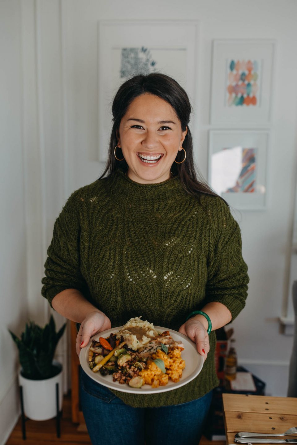Dark-haired woman in a green sweater holds a plate of carved turkey, in front of a festive tablescape & a full Thanksgiving dinner spread.