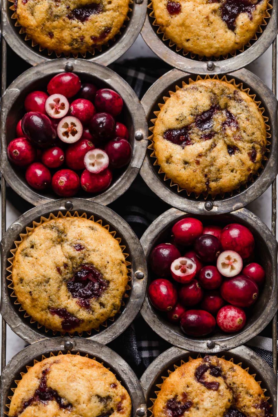 Cranberry corn muffins placed in a vintage muffin pan. 2 of the muffin cups are filled with fresh cranberries.
