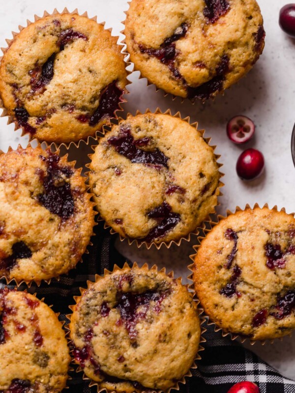 Swirled cranberry cornbread muffins shown on top of a black plaid napkin atop a white surface. Some fresh cranberries are scattered around the muffins.