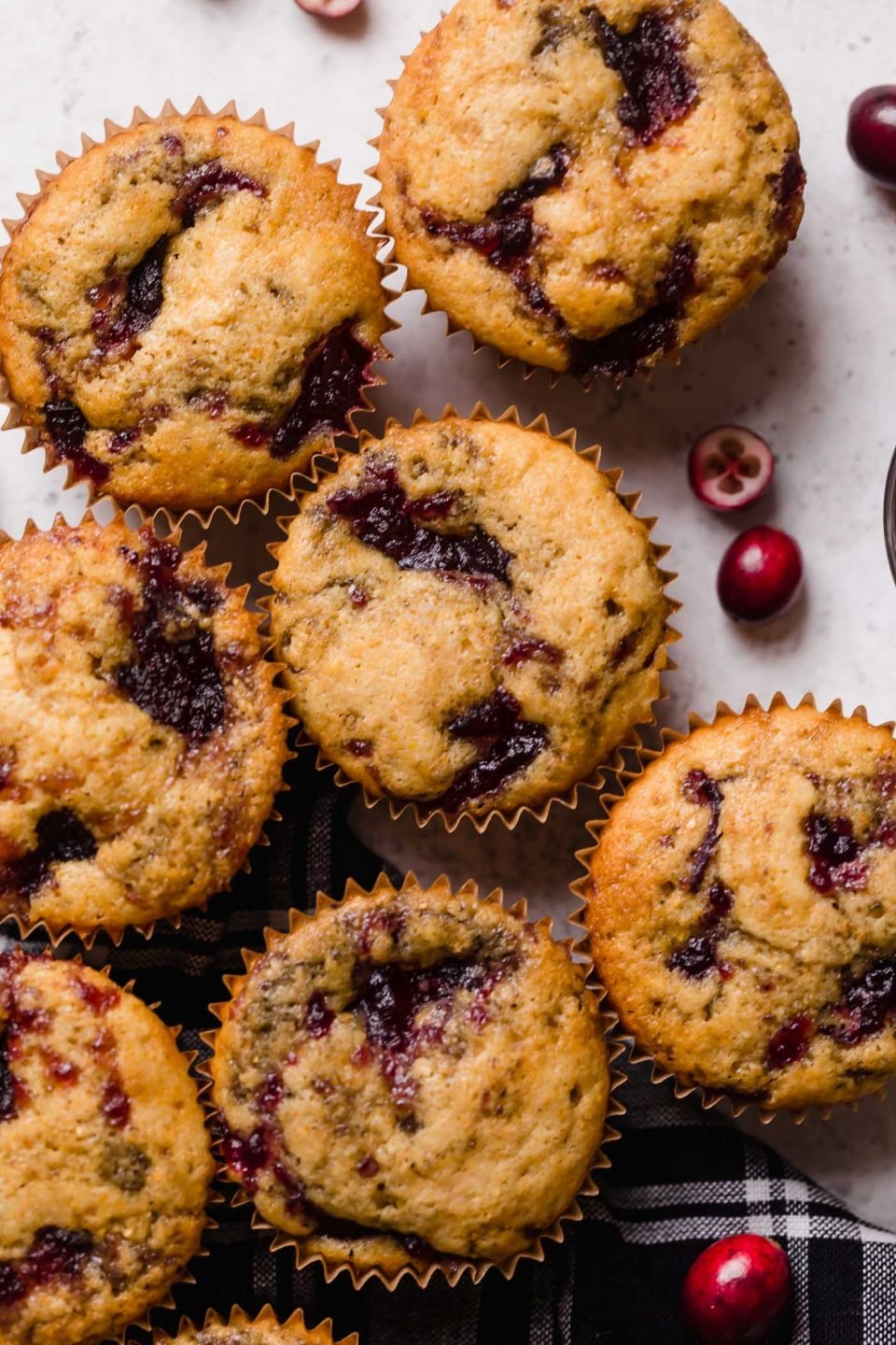 Swirled cranberry cornbread muffins shown on top of a black plaid napkin atop a white surface. Some fresh cranberries are scattered around the muffins.