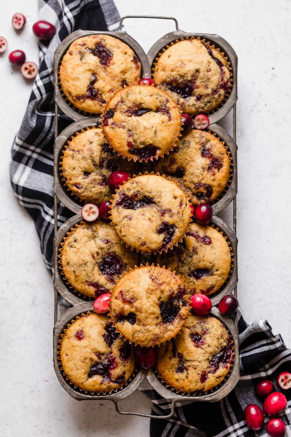 Cranberry cornbread muffins arranged in a vintage muffin pan. The muffin pan sits atop a black plaid linen napkin on a white surface, with some fresh cranberries scattered around it.