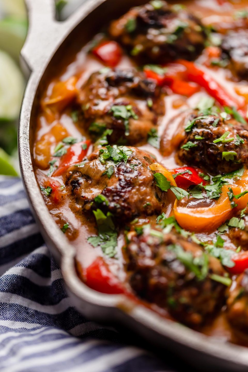A side angle shot of red curry meatballs fill a large cast iron skillet that sits atop a creamy white textured surface. The meatballs have been garnished with fresh chopped herbs. A blue and white striped linen napkin and lime wedges surround the skillet.