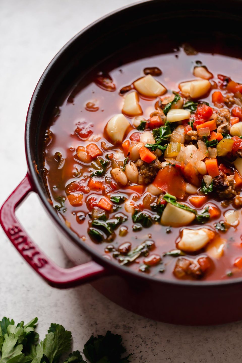 An side angle shot of a cherry red Staub cast iron dutch oven filled with a large batch of Hearty Minestrone Soup with Sausage. The dutch oven sits atop a creamy white textured surface. A bunch of fresh parsley rests alongside the dutch oven.