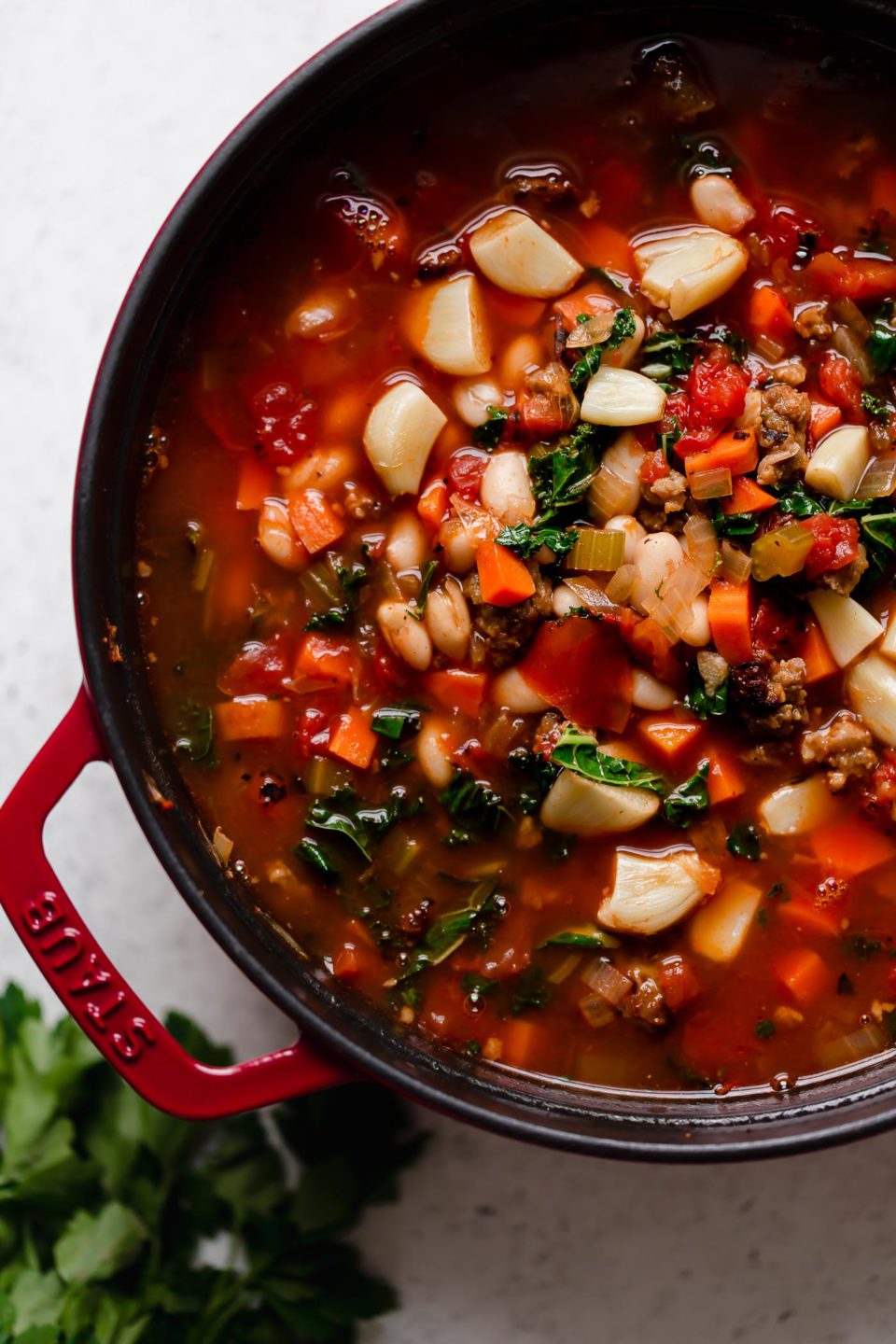 An overhead shot of a cherry red Staub cast iron dutch oven filled with a large batch of Hearty Minestrone Soup with Sausage. The dutch oven sits atop a creamy white textured surface. A bunch of fresh parsley rests alongside the dutch oven.