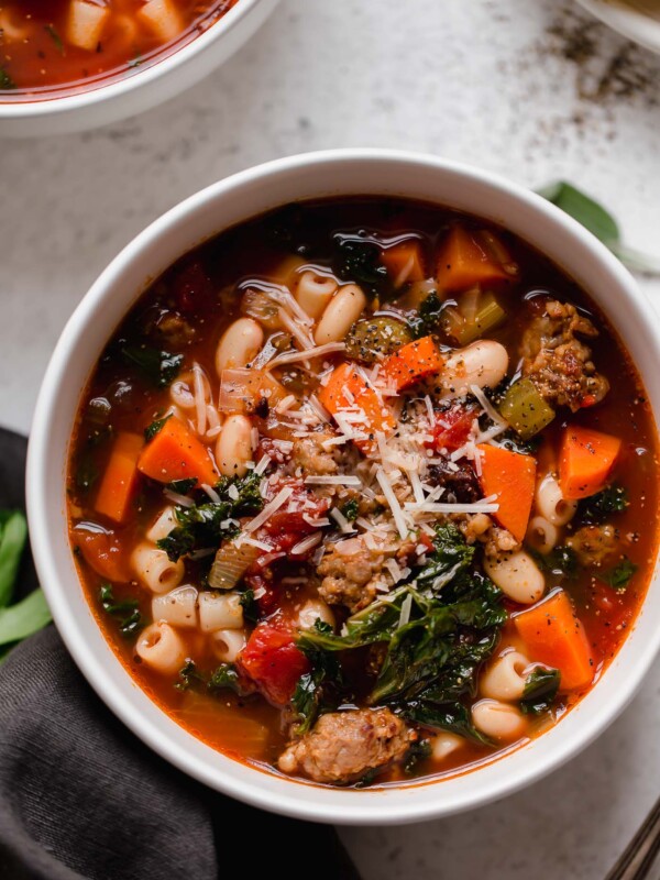 An overhead shot of a white soup bowl filled with Hearty Minestrone Soup with Sausage. The bowl sits atop a creamy white textured surface. The soup has been garnished with parmesan cheese and ground black pepper. A plate full of pieces of crusty bread, silverware, another bowl filled with minestrone soup, loose fresh herbs, and a dark grey linen napkin surround the bowl of soup at center.