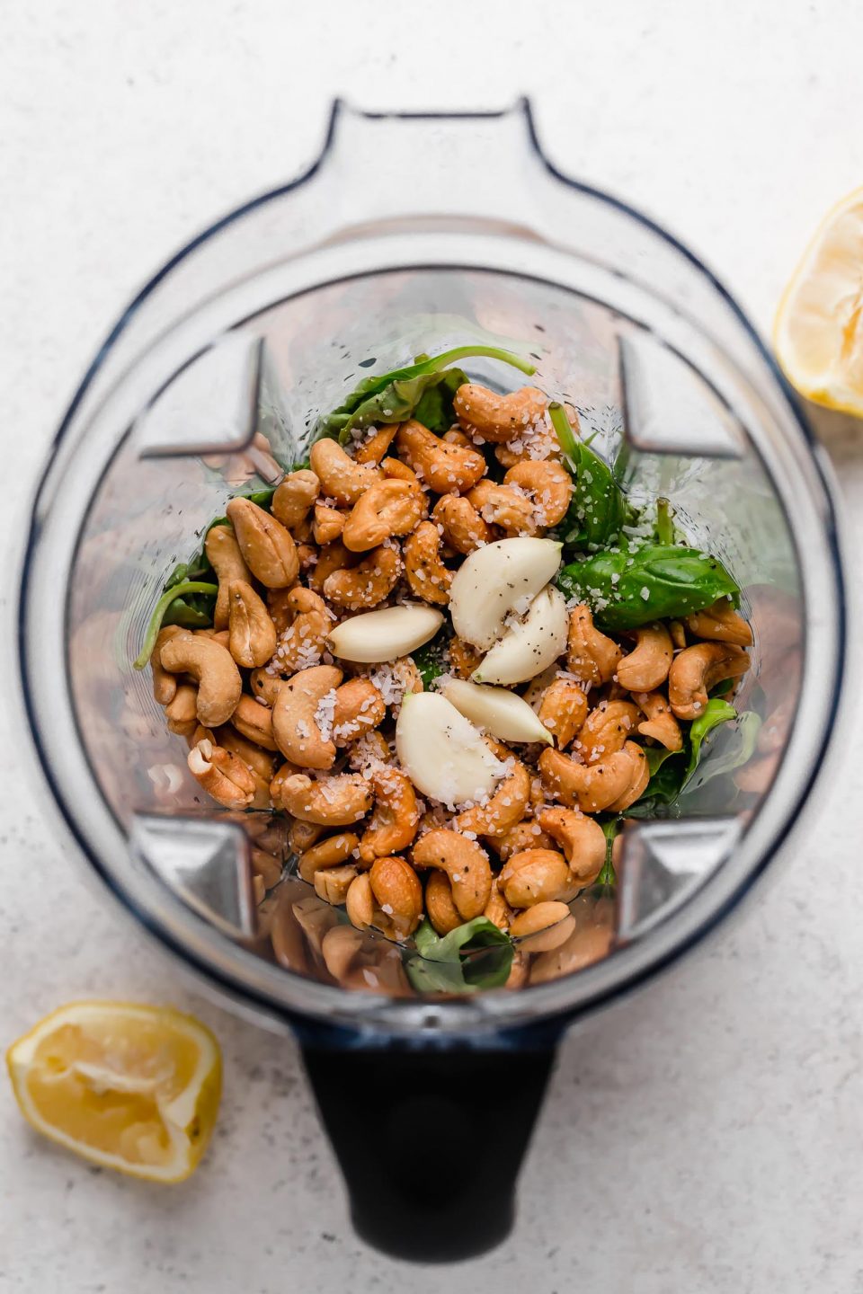 An overhead shot of cashews, garlic cloves, lemon juice, salt, and basil leaves in the bowl of a food processor atop a white surface. Two lemon wedges sit beside it.