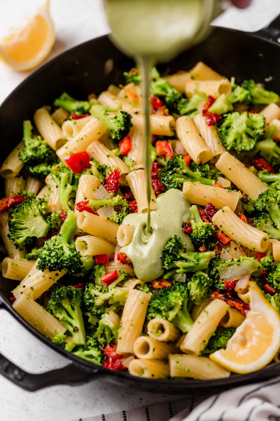 An overhead shot of vegan pasta sauce being poured over cooked pasta, sun-dried tomatoes, and cooked broccoli in a black skillet atop a white surface.