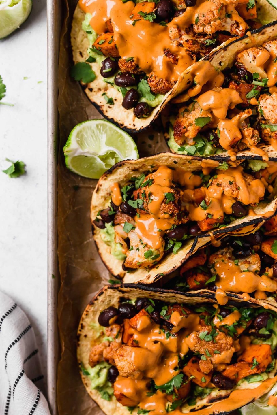 Vegetarian tacos made of roasted cauliflower, sweet potatoes & black beans arranged on a small rimmed baking sheet, topped with vegan cashew crema. 