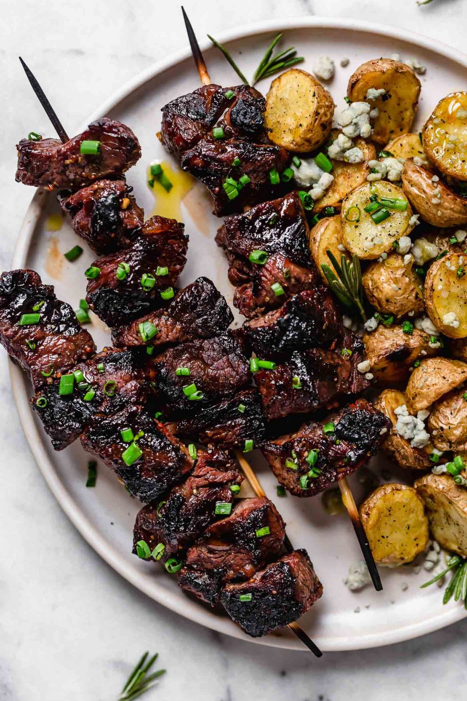 Sirloin steak kabobs are arranged atop a white plate alongside grilled baby potatoes. The meal has been garnished with fresh snipped chives and crumbled bleu cheese.
