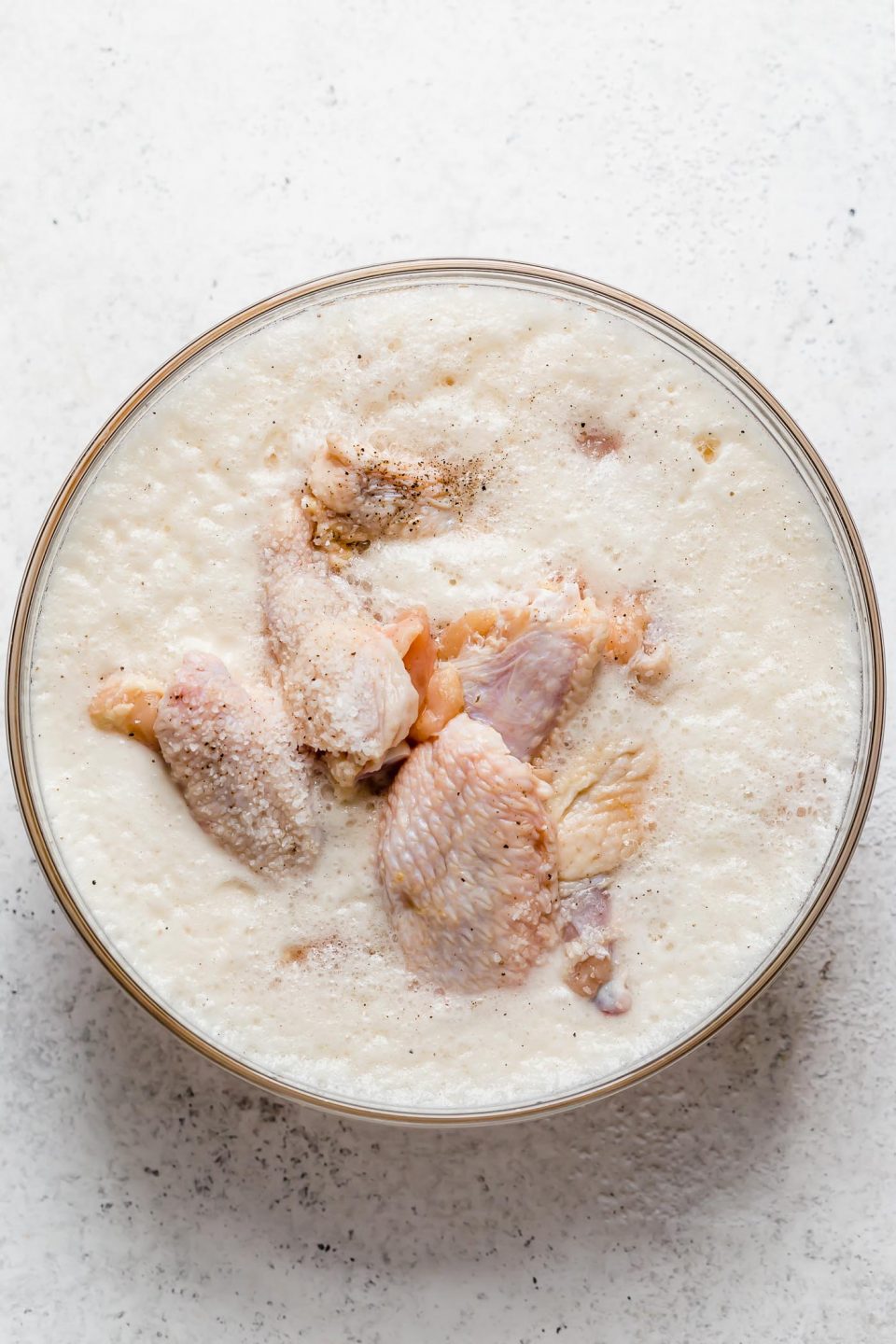 Chicken wings brine in beer inside of a clear glass mixing bowl that sits atop a creamy white textured surface.