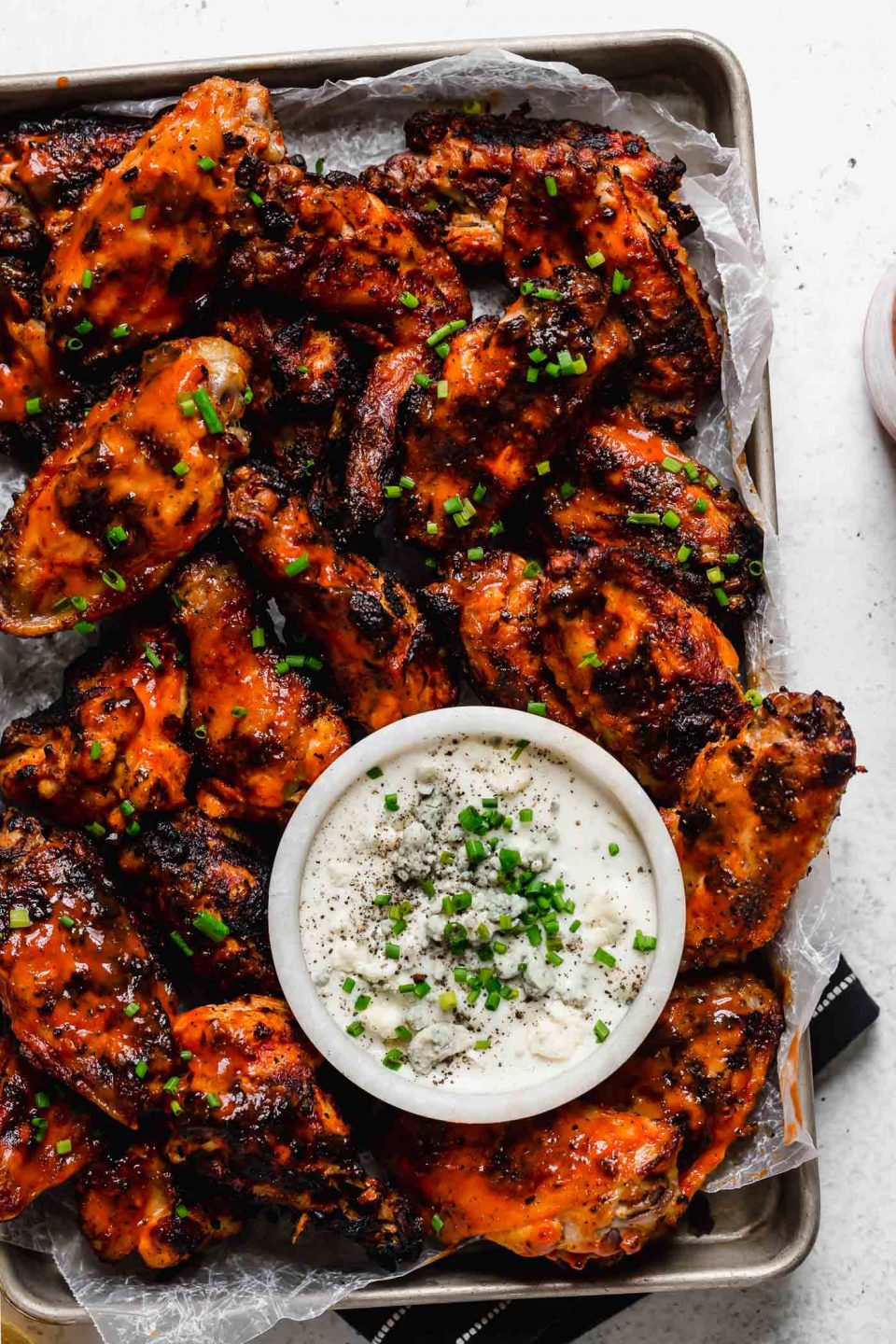 A small baking sheet of grilled chicken wings, & are topped generously with snipped chives. The tray sits atop a black & white striped linen on a white surface next to a bottle of Frank's RedHot hot sauce.