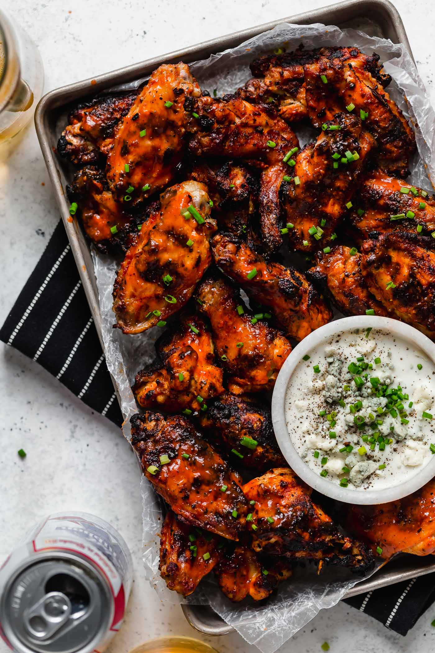 A small baking sheet of grilled chicken wings, topped generously with snipped chives & served with bleu cheese dressing. The tray sits atop a black & white striped linen on a white surface next to a can & a few glasses of PBR beer.