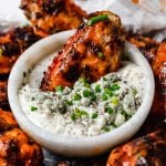 A crispy grilled wing dunked in a small bowl of bleu cheese dressing, in the center of a small baking sheet of grilled chicken wings, topped generously with snipped chives & served with bleu cheese dressing. The tray sits atop a black & white striped linen on a white surface next to a bottle of Franks RedHot hot sauce.