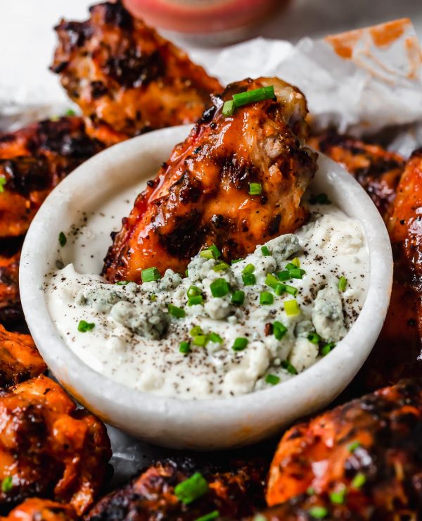 A crispy grilled wing dunked in a small bowl of bleu cheese dressing, in the center of a small baking sheet of grilled chicken wings, topped generously with snipped chives & served with bleu cheese dressing. The tray sits atop a black & white striped linen on a white surface next to a bottle of Franks RedHot hot sauce.