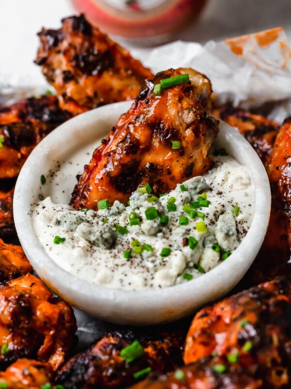 A crispy grilled wing dunked in a small bowl of bleu cheese dressing, in the center of a small baking sheet of grilled chicken wings, topped generously with snipped chives & served with bleu cheese dressing. The tray sits atop a black & white striped linen on a white surface next to a bottle of Franks RedHot hot sauce.