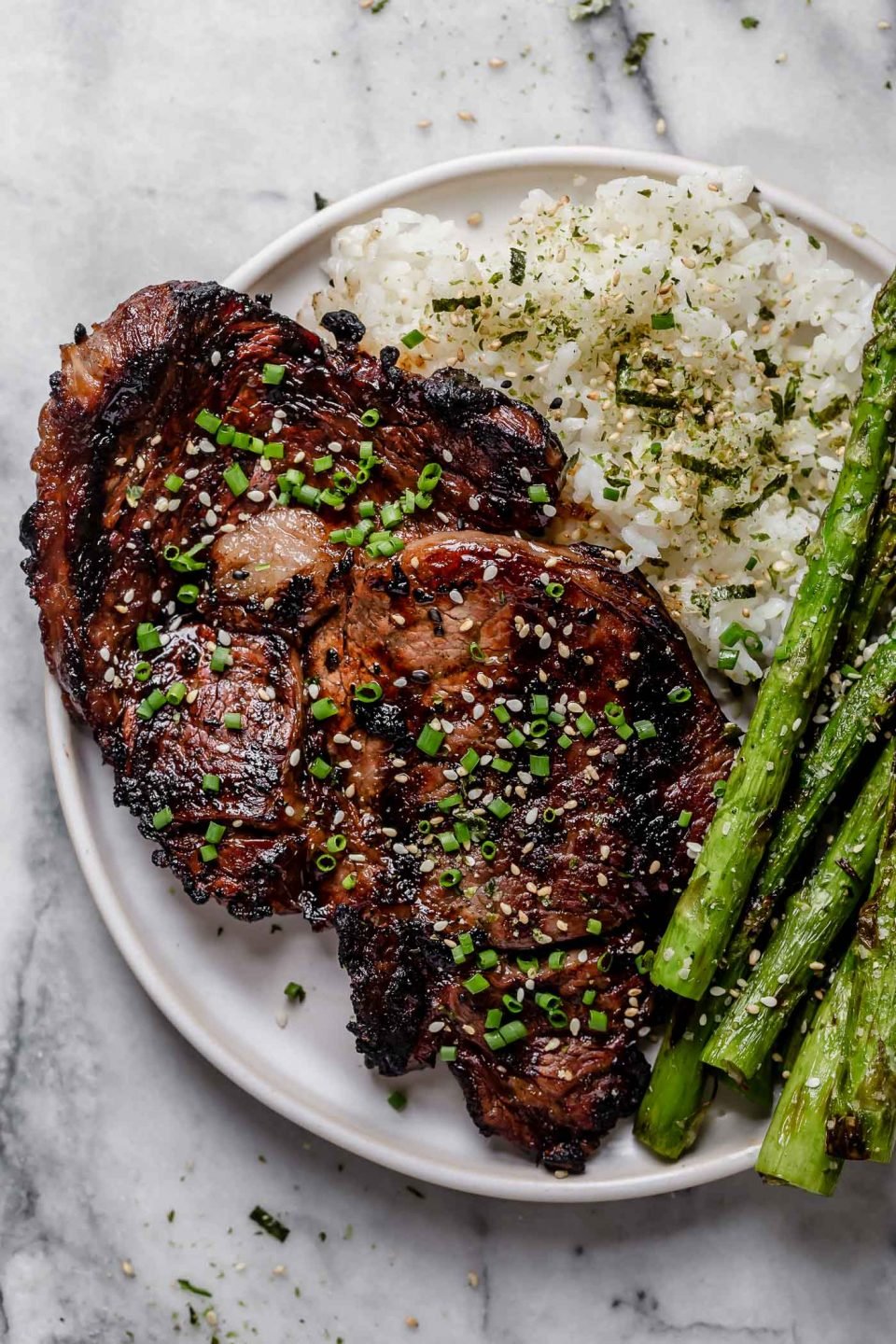 Grilled Teriyaki Ribeye Steaks shown on a white plate with seasoned sticky rice & grilled asparagus sits atop a gray and white marble surface. The ribeye has been marinated teriyaki & is garnished with freshly snipped chives & sesame seeds.