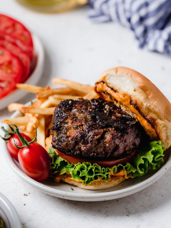bacon cheddar stuffed burger served on a bun with french fries next to another plated burger, plated burger toppings & a ice cold beer.