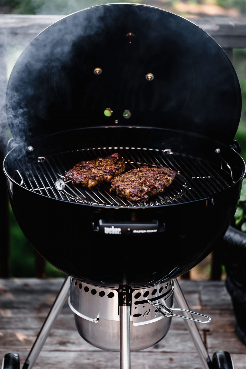 Two steaks lay on top of an open charcoal grill with a thermometer sticking in one of the steaks to check the internal temperature