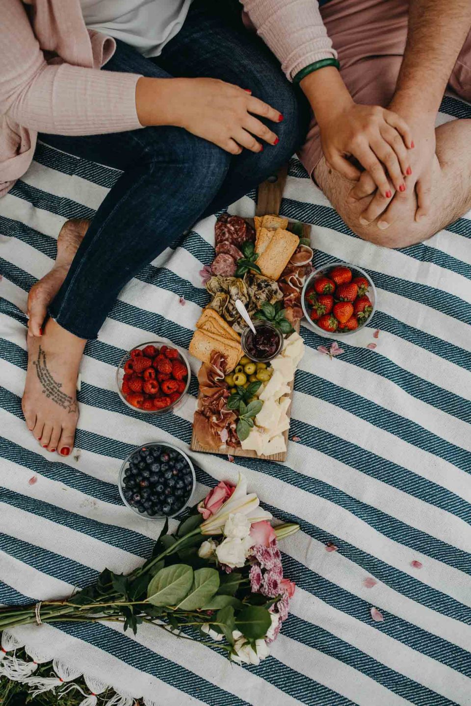 overhead view of a couple seated on a blue striped picnic blanket on the grass next to a lake with a summer picnic charcuterie board for 2, along with fresh flowers & a picnic basket.