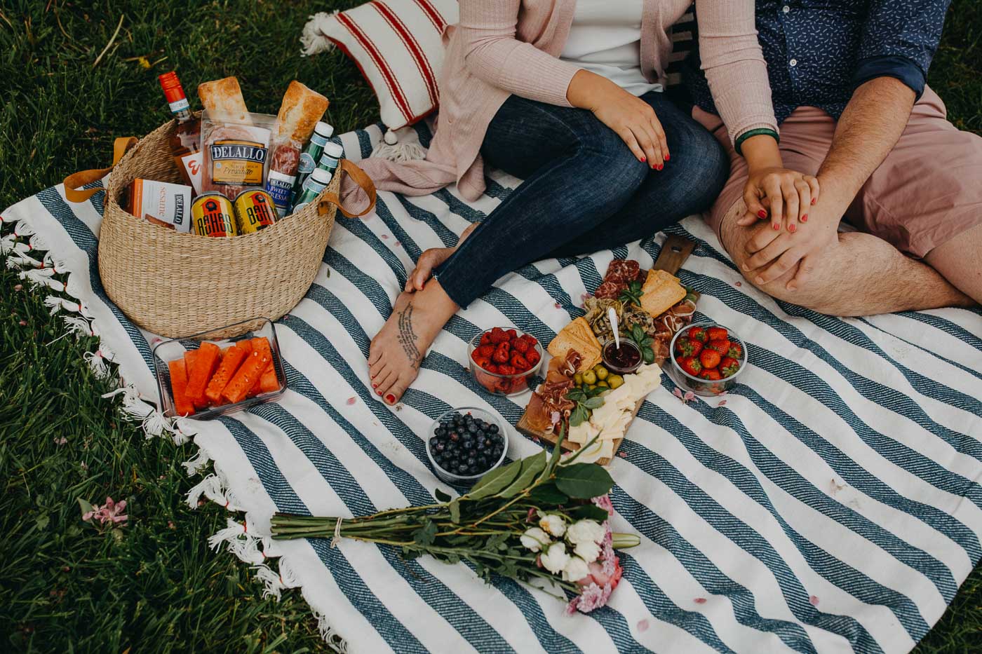 overhead view of a couple seated on a blue striped picnic blanket on the grass next to a lake with a summer picnic charcuterie board for 2, along with fresh flowers & a picnic basket. 