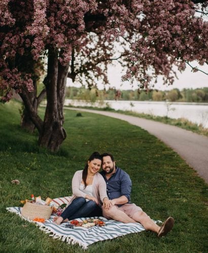 a couple seated on a blue striped picnic blanket on the grass next to a lake with a summer picnic charcuterie board for 2.