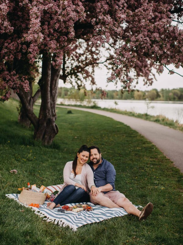 a couple seated on a blue striped picnic blanket on the grass next to a lake with a summer picnic charcuterie board for 2.