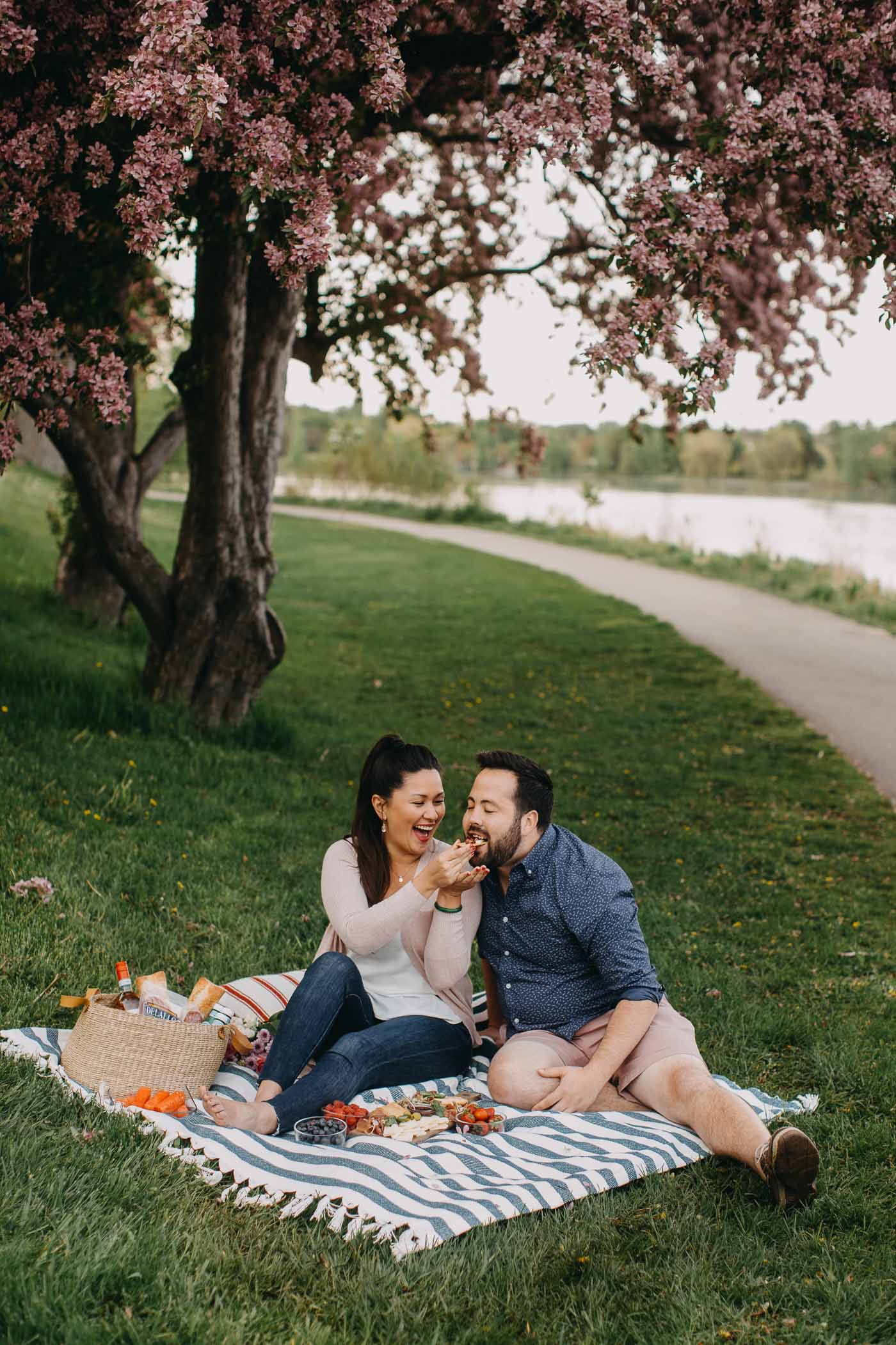 a couple seated on a blue striped picnic blanket on the grass next to a lake with a summer picnic charcuterie board for 2. she is feeding him a bite of charcuterie & they are both laughing. 