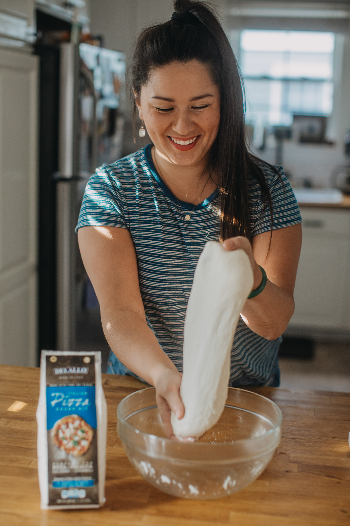 Woman in blue shirt pulling homemade pizza dough out of clear glass bowl. Bowl is sitting next to DeLallo pizza dough kit package.