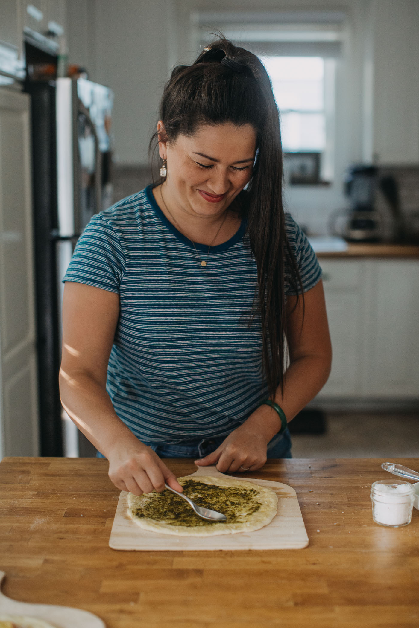 woman in blue shirt showing how to make homemade pizza for pizza night by spreading fresh pesto on pizza crust.